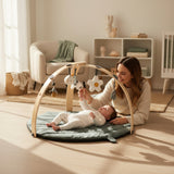 Baby lying on a green play mat with a wooden arch and hanging toys on a white background