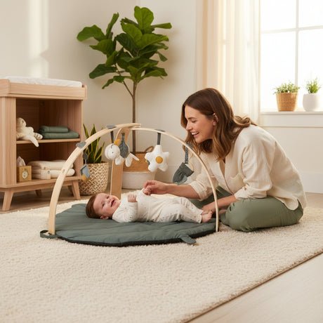 Baby lying on a green play mat with a wooden arch and hanging toys on a white background