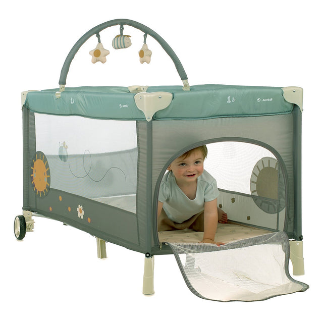 Child playing inside a green and beige playpen with toys hanging above.